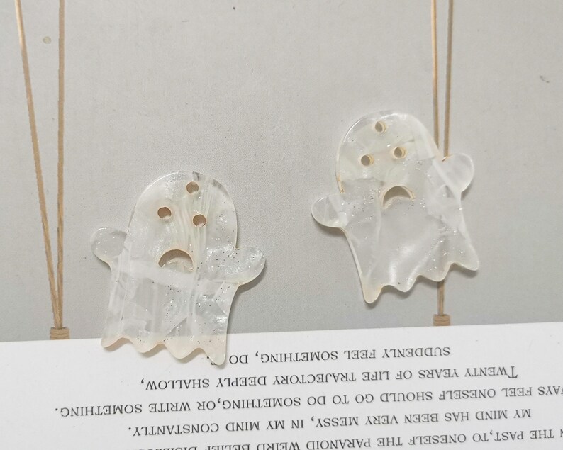 Puede incluir: Dos adornos blancos en forma de fantasma con detalles de purpurina. Los fantasmas tienen una cara triste con tres agujeros para los ojos y una boca.