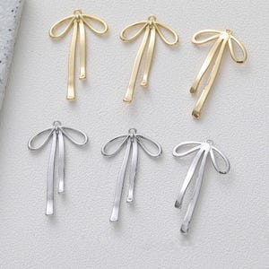 Könnte beinhalten: Sechs goldene und silberne Anhänger in Form einer Schleife mit einer herabhängenden Schlaufe. Die Anhänger sind perfekt für die Schmuckherstellung oder um handwerklichen Arbeiten einen Hauch von Eleganz zu verleihen.