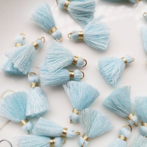May include: A collection of light blue tassels with gold accents. The tassels are arranged on a white surface.