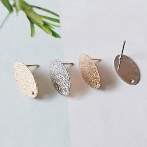Könnte beinhalten: Drei ovale Ohrringrohlinge mit strukturierten Oberflächen in Gold, Silber und gebürstetem Silber. Die Ohrringrohlinge haben einen Stift und ein Loch zum Anbringen von Ohrringbefestigen.