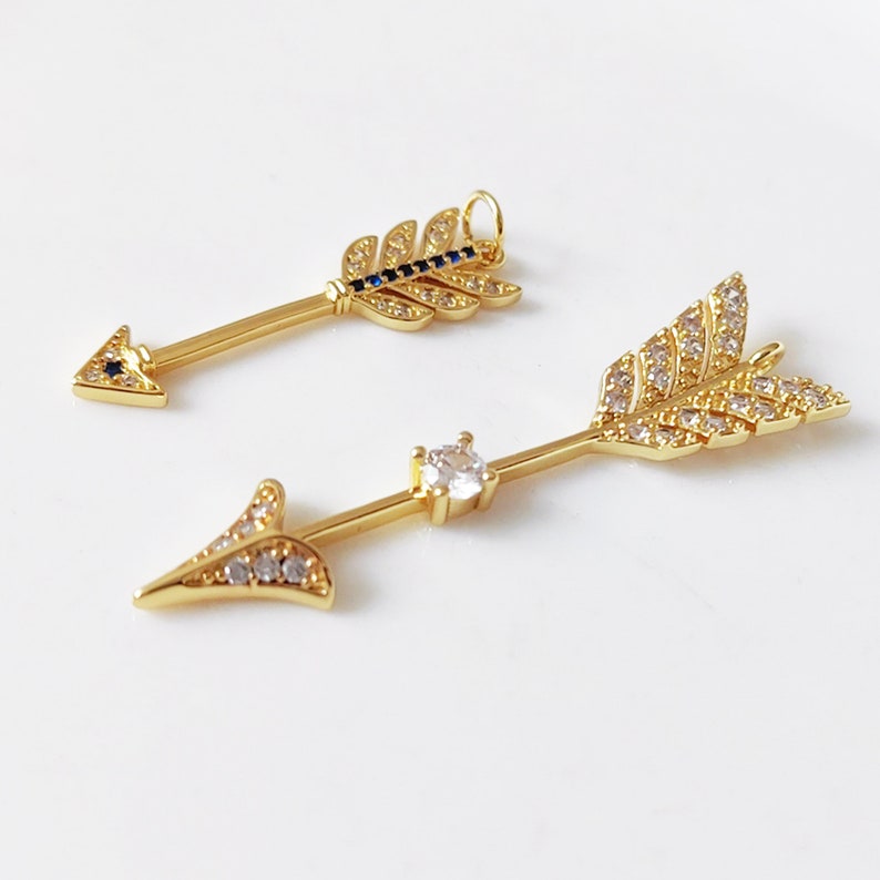 May include: Two gold arrow charms with clear and blue crystal accents. The arrowheads are pointed and the feathers are detailed.