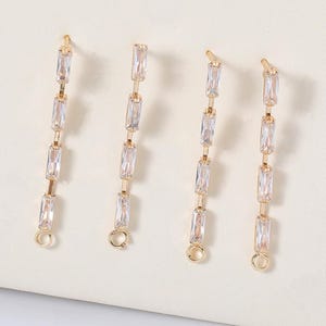 May include: Four gold-toned, linear earrings with rectangular, clear crystals. Each earring features a series of crystals connected by gold links, with a small loop at the bottom for attachment. The earrings are displayed on a light-colored surface.