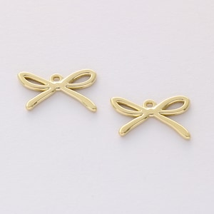 Pode incluir: Dois pingentes dourados em forma de laço. Cada pingente tem uma argola na parte superior para prender em joias. Os laços têm um design simples e elegante, com linhas curvas e um acabamento polido. Os pingentes estão em um fundo branco liso.