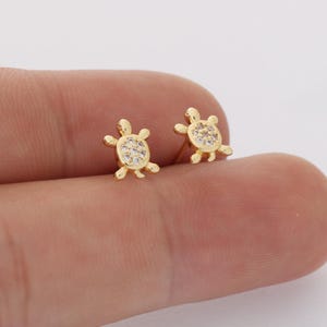 以下が含まれることがあります： カメの形をしたゴールドトーンのスタッドピアス一対。それぞれのカメの甲羅には、小さく透明な輝く石が埋め込まれています。ピアスは光沢のある金属仕上げで、4本の小さな足と頭部がデザインされています。