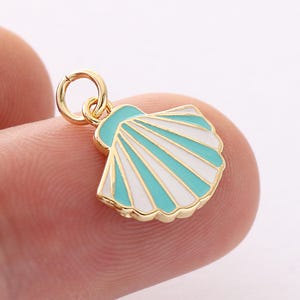 Puede incluir: Un dije de concha dorada con un diseño de esmalte azul claro y blanco. El dije tiene un pequeño lazo para sujetarlo a un collar o pulsera. El diseño de la concha es detallado y llamativo.