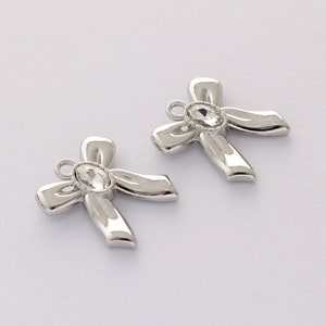 Op de afbeelding: Twee zilveren strikbedels met heldere strasssteentjes in het midden. De strikken zijn in een knoop geknoopt en hebben bovenaan een lus om aan een ketting of armband te bevestigen.