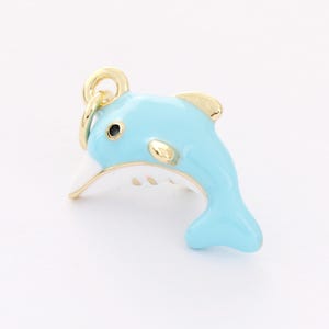 May include: A gold-toned charm in the shape of a dolphin. The dolphin is blue with a white belly and has a gold-toned ring at the top for attaching to a necklace or bracelet.