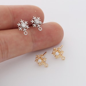 Pode incluir: Dois pares de brincos de pino em forma de floco de neve, um par em tom prateado e outro em tom dourado. Cada floco de neve apresenta uma pedra transparente central e um pequeno anel circular na base para fixar componentes de joalharia.