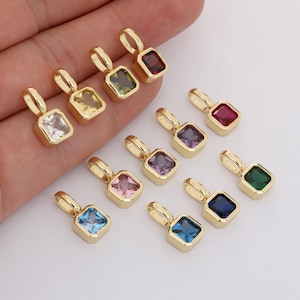 Könnte beinhalten: Eine Auswahl an goldfarbenen Anhängern, jeweils mit einem quadratischen Edelstein in verschiedenen Farben. Die Edelsteine sind weiß, gelb, grün, rot, lila, rosa, blau und dunkelblau. Die Anhänger haben eine kleine Schlaufe.