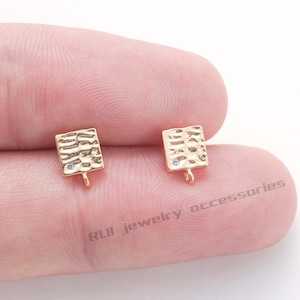 May include: Two gold-colored, textured square jewelry findings with a small, clear gem and a loop at the bottom. The findings are for jewelry making and are shown against a light background.