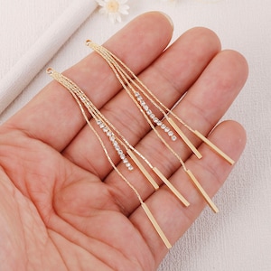 Op de afbeelding: Een paar goudkleurige oorbellen met een bungelend ontwerp. Elke oorbel heeft meerdere strengen van een fijne ketting, waarvan sommige versierd zijn met kleine, sprankelende kristallen. Rechthoekige gouden staven hangen aan de onderkant van elke oorbel.