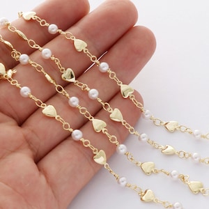 May include: Four gold chains with small heart charms and white pearl beads. The chains are layered on top of each other and are displayed on a hand.