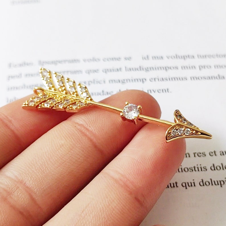 May include: Gold arrow charm with clear crystal accents. The arrow has a feather design on one end and a pointed tip on the other.