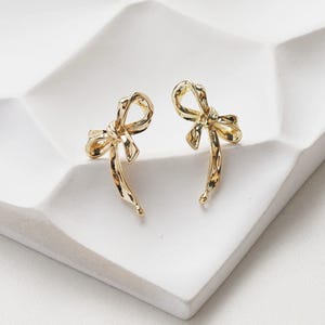 May include: A pair of gold bow-shaped earrings. The earrings are made of a textured metal and have a simple, elegant design.