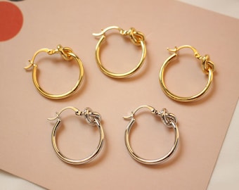 May include: Five hoop earrings in gold and silver tones. The earrings feature a knot design at the top of the hoop. The gold earrings are in the majority, with two silver earrings at the bottom.