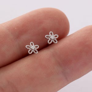 May include: Two small, silver-colored flower stud earrings. Each earring features a five-petal design with a small, clear stone in the center. The earrings are shown against a skin-toned background.