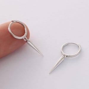 May include: A pair of silver hoop earrings with a pointed silver dangle. The earrings are shown on a finger.