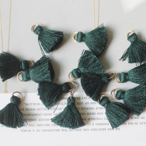May include: Ten dark green tassel charms with gold jump rings. The tassels are made of soft yarn and have a fluffy texture.