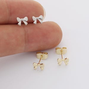 May include: A pair of silver-toned bow-shaped stud earrings and a pair of gold-toned bow-shaped stud earrings. Each earring features a small, delicate bow design with a post back and a matching metal butterfly clutch closure for secure fastening.