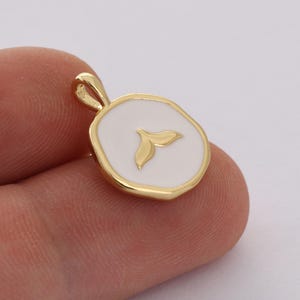 Könnte beinhalten: Goldener Anhänger mit einem weißen Emaille-Kreis, der ein goldenes Wal-Schwanz-Design zeigt.