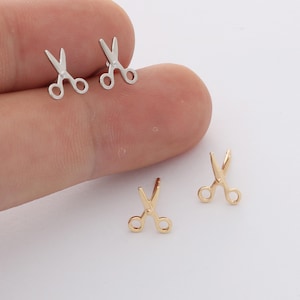 May include: A pair of silver-toned metal stud earrings shaped like small scissors and a pair of gold-toned metal stud earrings shaped like small scissors. Each earring features two circular finger loops and pointed blades in a simple, clean design.