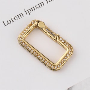 May include: A gold-coloured rectangular clasp, encrusted with small, clear gemstones. The clasp has a hinged design for opening and closing. The background is a white surface with faint text.