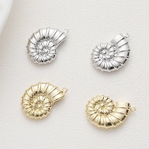 Pode incluir: Quatro pingentes de concha de nautilus prateados e dourados com um laço para prender em joias. Os pingentes são detalhados com linhas em relevo e um design em espiral.