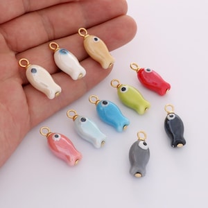 May include: A selection of colourful ceramic fish charms with gold-coloured accents. Each charm has a small eye and a loop for attaching to jewellery or other crafts. The fish are in various colours, including red, blue, green, and beige.