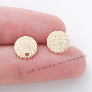 Op de afbeelding: Twee gouden metalen cirkels met een klein gaatje in het midden. De cirkels hebben een getextureerd oppervlak.