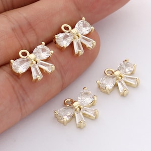 Op de afbeelding: Gouden strikvormige bedels met heldere, sprankelende cubic zirconia stenen. Elke bedel heeft een centrale peervormige steen en rechthoekige accenten, perfect voor het maken van sieraden. De bedels hebben een kleine lus.
