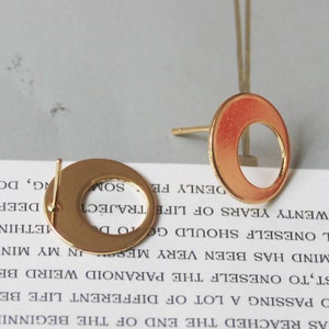 May include: Two gold-colored, open-circle stud earrings. The earrings have a post for insertion and a circular design with a hollow center. The earrings are displayed on a light-colored surface.