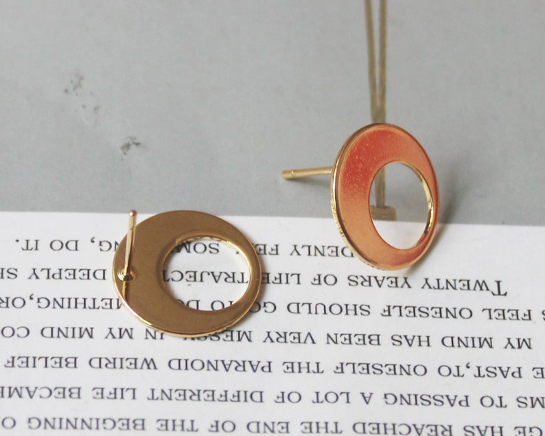 May include: Two gold-colored, open-circle stud earrings. The earrings have a post for insertion and a circular design with a hollow center. The earrings are displayed on a light-colored surface.