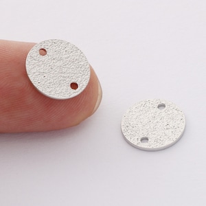 May include: Two silver-toned metal round disc charms with two holes each. The charms have a textured surface.