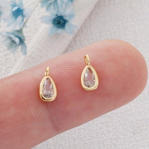 May include: Two gold teardrop-shaped charms with clear crystal accents. The charms are on a finger.
