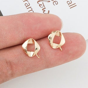 May include: Two small, gold-colored earrings. Each earring has a square, open-frame design with a textured surface. A small post extends from the bottom of each earring. The earrings are displayed on a light background.