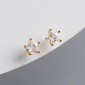 May include: A pair of gold-coloured star-shaped stud earrings. Each earring features a clear, square-cut gemstone at the centre, with gold prongs forming the star points. The earrings are set against a white background.