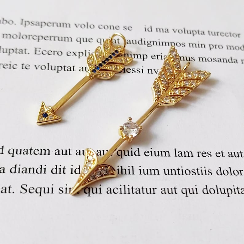 May include: Two gold arrow charms with clear and blue rhinestones. The arrowheads are pointed and the feathers are detailed.