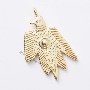 Peut inclure: Pendentif en or ton sur ton représentant un oiseau stylisé avec un petit strass transparent sur la tête et un plus gros strass rond au centre. Le pendentif a une boucle en haut pour l'attacher à une chaîne.