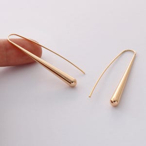 Op de afbeelding: Een paar goudkleurige oorhangers. Elke oorbel heeft een lange, taps toelopende, druppelvormige hanger met een afgeronde bodem. De oorbellen hebben een eenvoudig, gebogen draadontwerp voor het oor. De oorbellen worden op een witte achtergrond getoond.