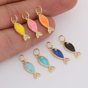 May include: A selection of fish-shaped charms in various colours. Each charm has a gold-coloured frame, a brightly coloured enamel body, and a crystal-studded tail. The charms are in yellow, pink, orange, blue, and black.