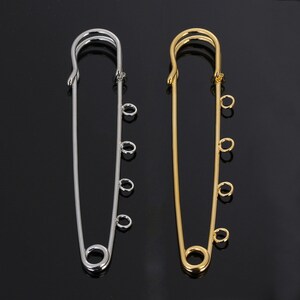 May include: Two safety pins with decorative loops. The safety pin on the left is silver and the safety pin on the right is gold.