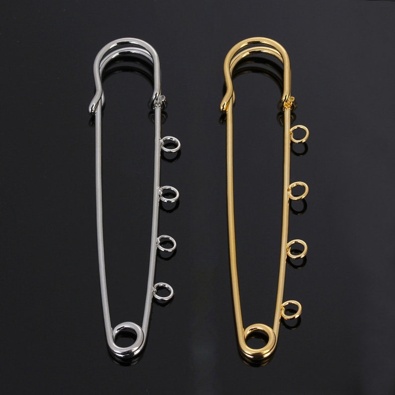 May include: Two safety pins with decorative loops. The safety pin on the left is silver and the safety pin on the right is gold.