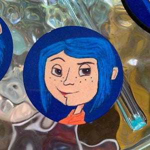 May include: A round blue and white sticker with a cartoon illustration of a girl with blue hair and freckles. She is wearing an orange shirt.