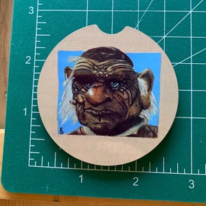 Op de afbeelding: Een ronde onderzetter met een bruine achtergrond en een vierkant beeld van een cartoonfiguur met een grote neus en wit haar. Het personage heeft een blauwe achtergrond en draagt een bruin shirt.