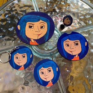 May include: Set of four round coasters featuring a cartoon character with blue hair and a red dress. The coasters are made of wood and have a dark blue background. The character is from the movie "Coraline".