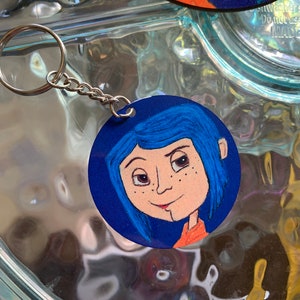 May include: A round blue and orange key chain featuring a cartoon illustration of a girl with blue hair and freckles. The girl is wearing a red and orange shirt.