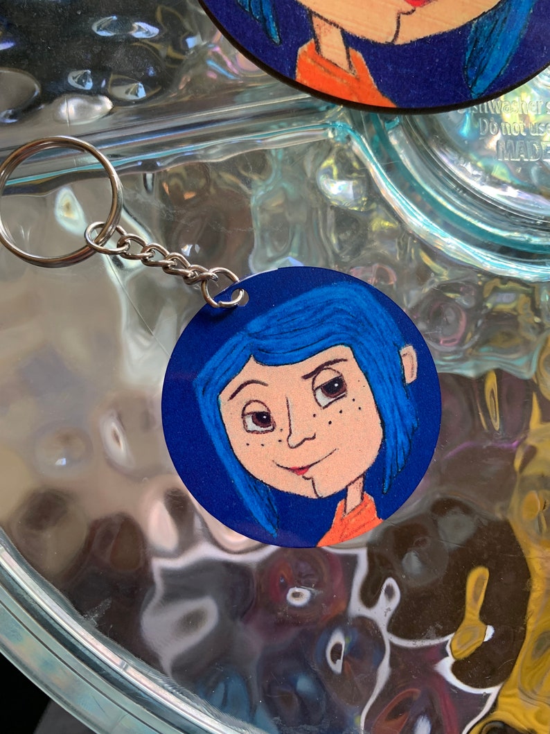 May include: A round blue and orange key chain featuring a cartoon illustration of a girl with blue hair and freckles. The girl is wearing a red and orange shirt.
