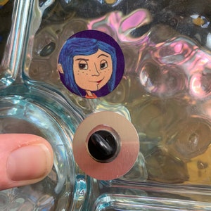 May include: A close-up of a glass surface with a round sticker featuring a cartoon girl with blue hair and a purple background. A silver metal drain stopper is visible below the sticker.