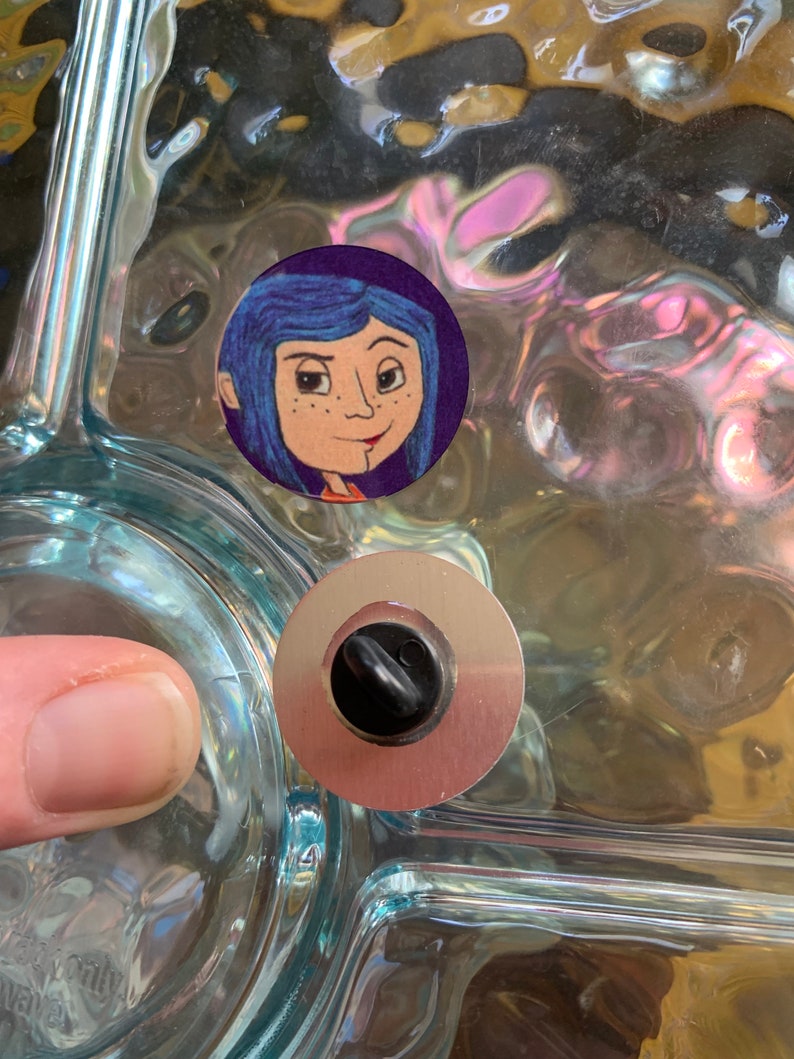 May include: A close-up of a glass surface with a round sticker featuring a cartoon girl with blue hair and a purple background. A silver metal drain stopper is visible below the sticker.