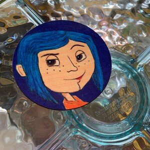 May include: A hand-painted wooden round coaster featuring a cartoon portrait of a girl with blue hair and a red shirt. The background is a dark blue color.